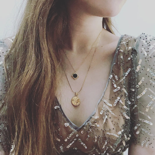 A woman wearing a gold-colored necklace with a pendant featuring a coin-shaped charm.